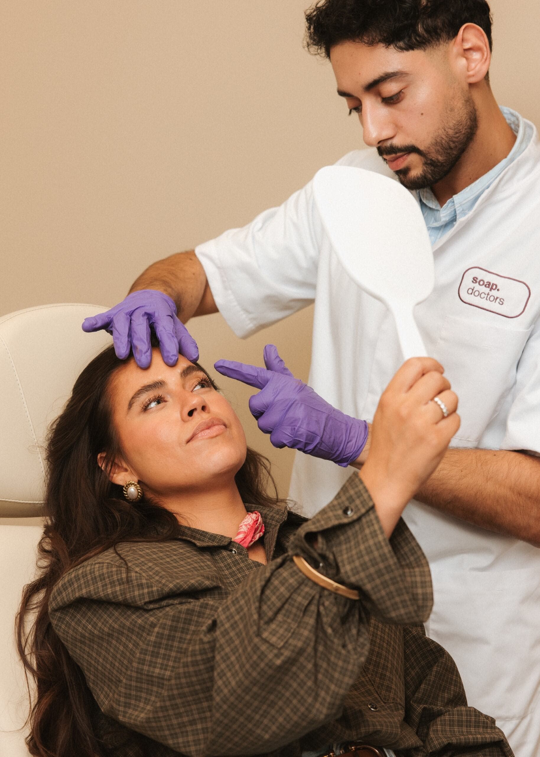 A woman in a clinic holds a mirror, examining her reflection, while a professional in a white coat adjusts her forehead, wearing purple gloves.